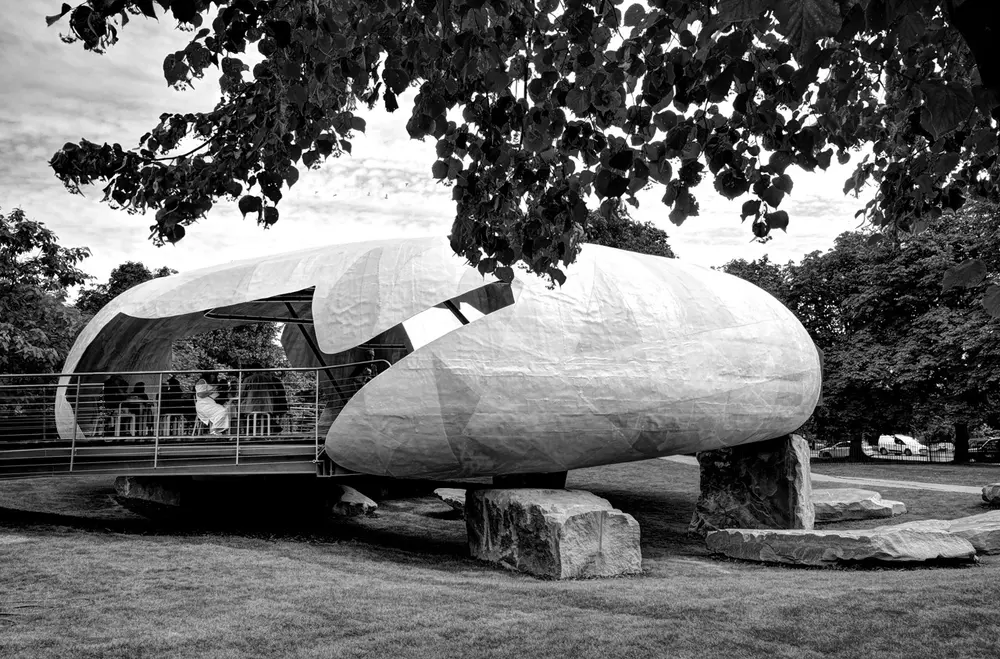 A black and white photo of a pavilion like a cracked, hollow white shell, resting on large rough boulders on a lawn under trees.