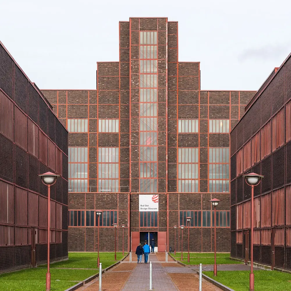 A tall, symmetrical brick and steel industrial building with long vertical windows, at the end of a paved path between two long brick wings.