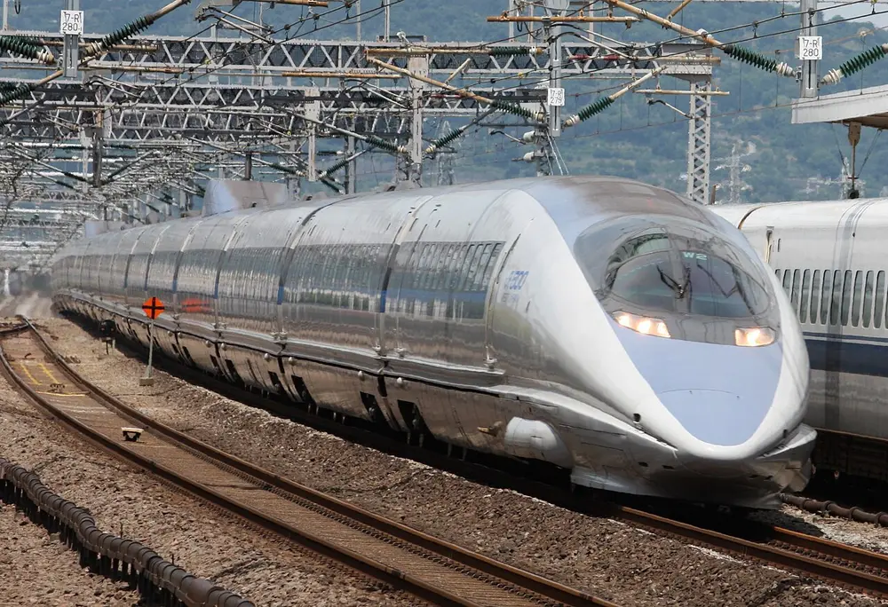 A silver bullet train with a very long, smooth, pointed nose and a round cab window, running on tracks under overhead wires.