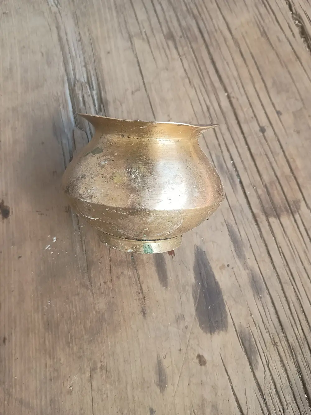 A small, round brass lota with a flared rim, standing on a weathered wooden table.