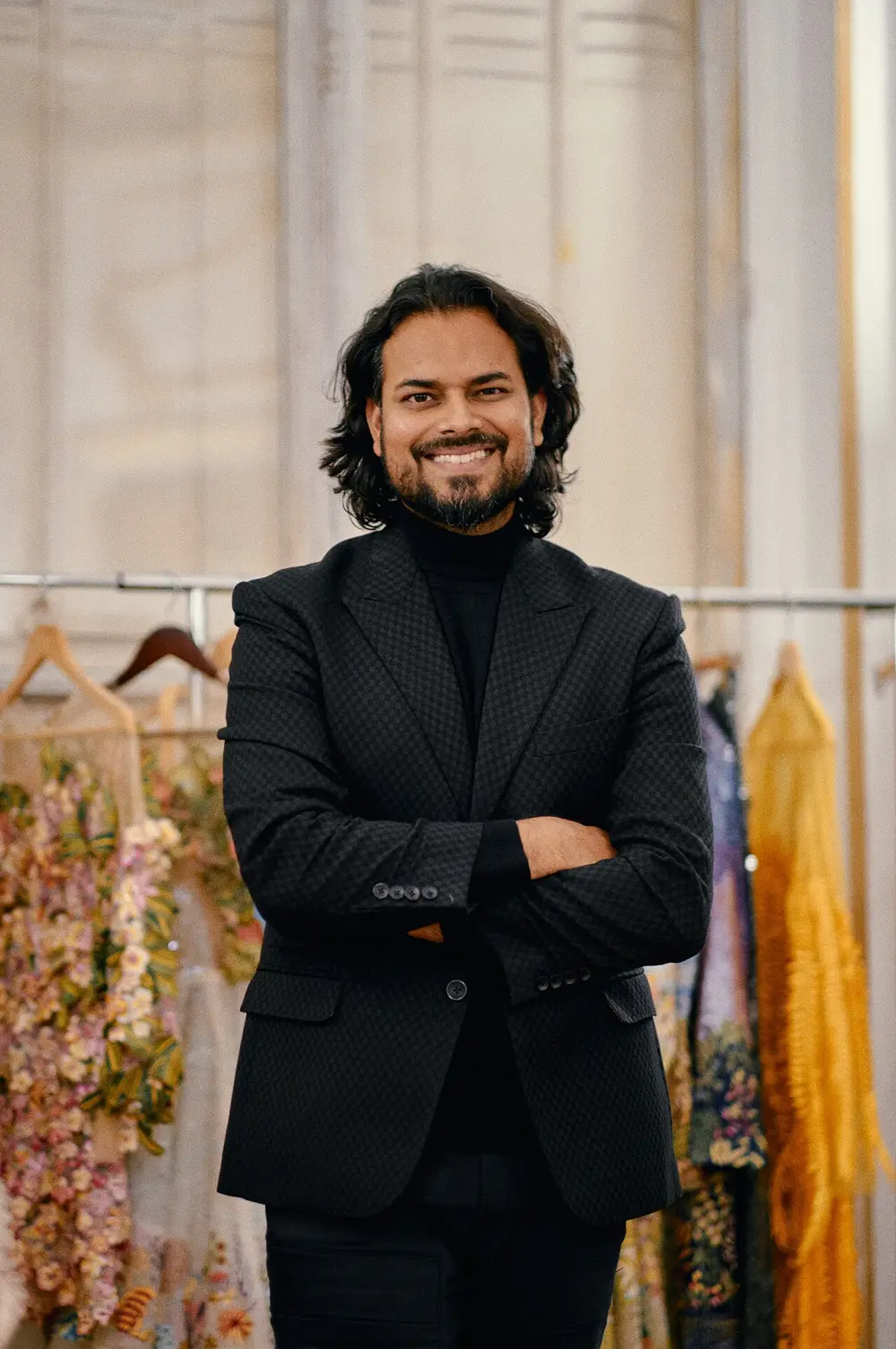The designer Rahul Mishra smiling, arms folded, in a black jacket, in front of rails of embroidered dresses.