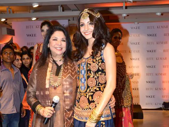 The designer Ritu Kumar, on the left in a brown printed outfit and holding a microphone, beside the actor Anushka Sharma at a store launch.