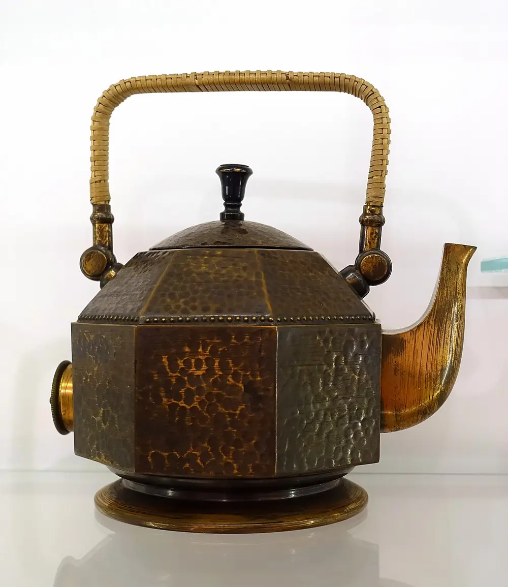 An eight-sided electric kettle of hammered copper with a domed lid, a black knob, a curved spout and a cane-wrapped handle.