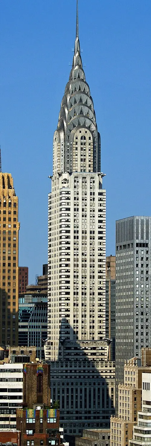 A tall white skyscraper with a shiny steel crown of stacked arches pierced by triangular windows, rising to a thin spire against a blue sky.
