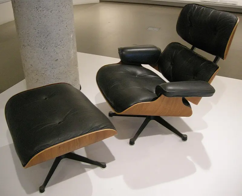 A low lounge chair and matching footstool, each made of curved wooden plywood shells holding black leather cushions, on black swivel bases.