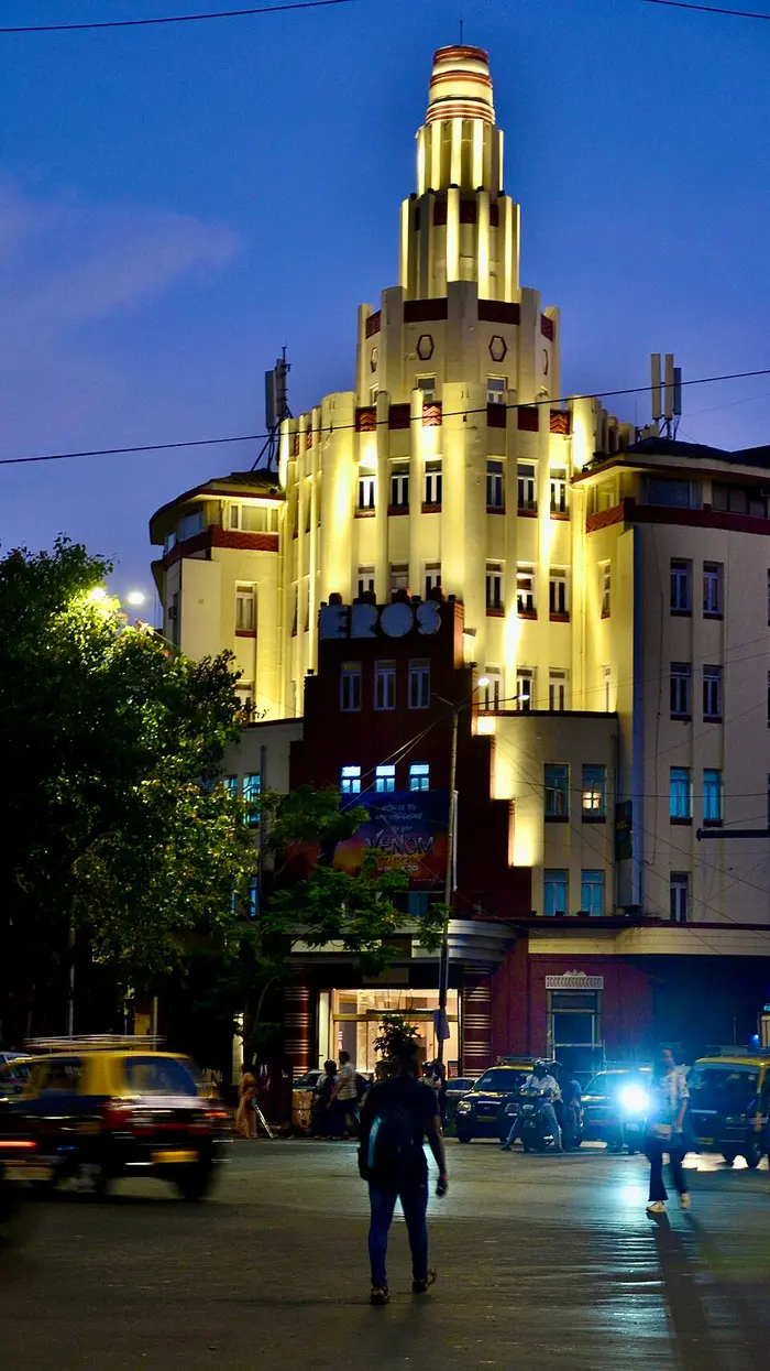 A cream building lit up at dusk, with a stepped central tower, red bands, rounded corners and the word EROS above the entrance, on a busy Mumbai street with taxis.