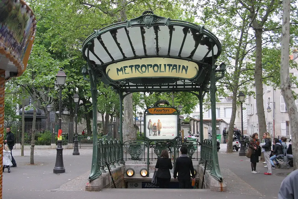 A green cast-iron Métro entrance with a fan-shaped glass canopy, curving plant-like posts, two round lamps and a sign reading METROPOLITAIN, in a tree-lined Paris square.