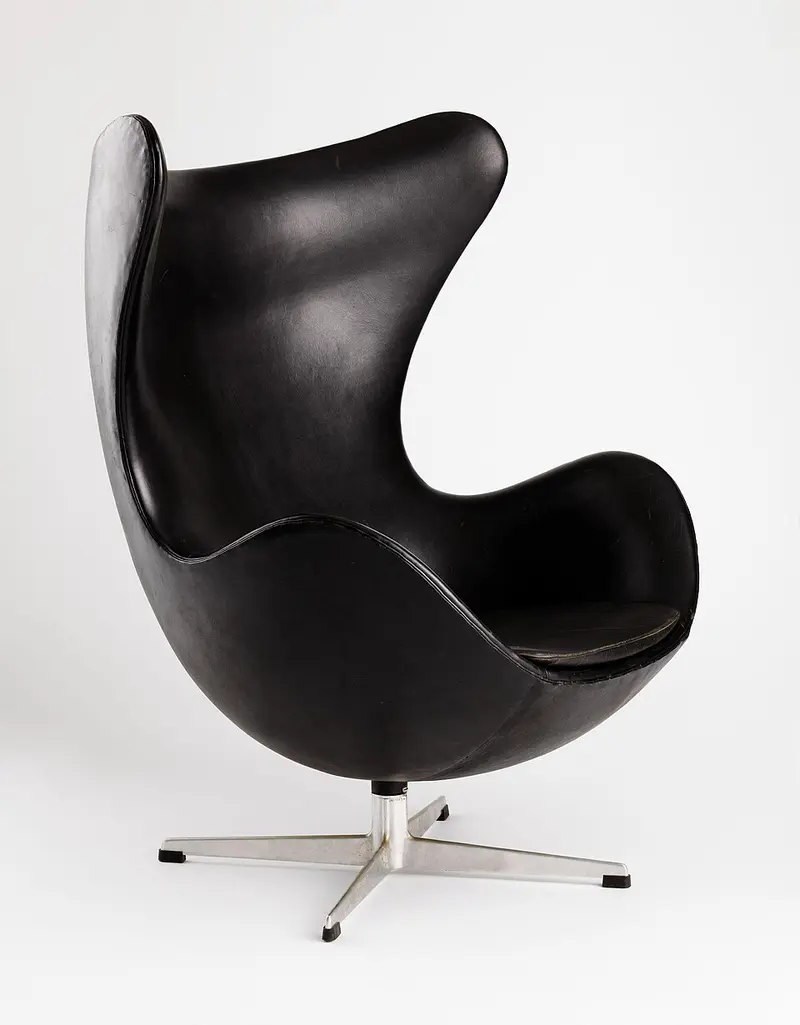 A black leather armchair shaped like an open egg shell, with a high curved back and wings, on a four-pronged metal swivel base.