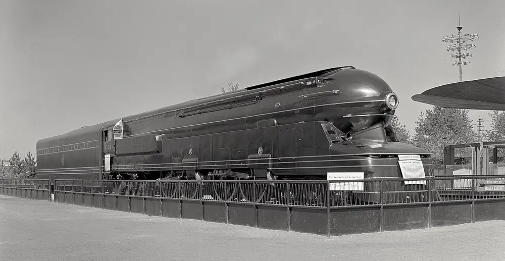 A black and white photo of a huge steam locomotive covered in a smooth, rounded steel shell with a bullet-shaped nose and long horizontal lines, on display behind a fence.