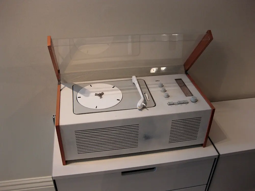 A white metal radio and record player with light wooden side panels and a raised clear plastic lid, showing a white turntable and a neat row of small grey buttons and knobs.