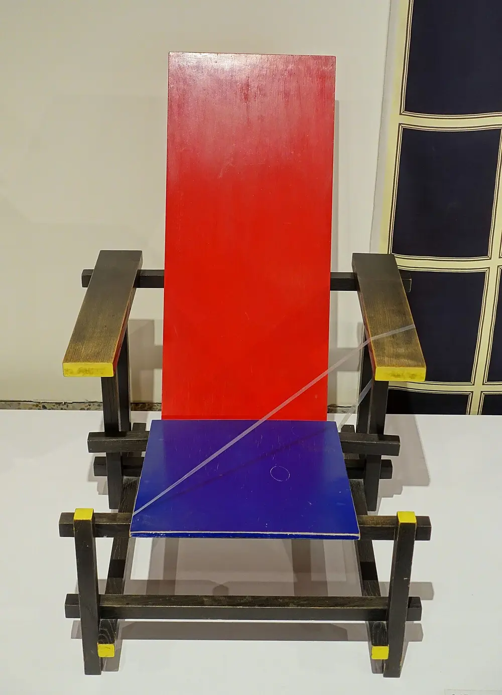 An armchair made of flat boards and square wooden rails, with a red back, a blue seat, and black rails with yellow-painted ends.