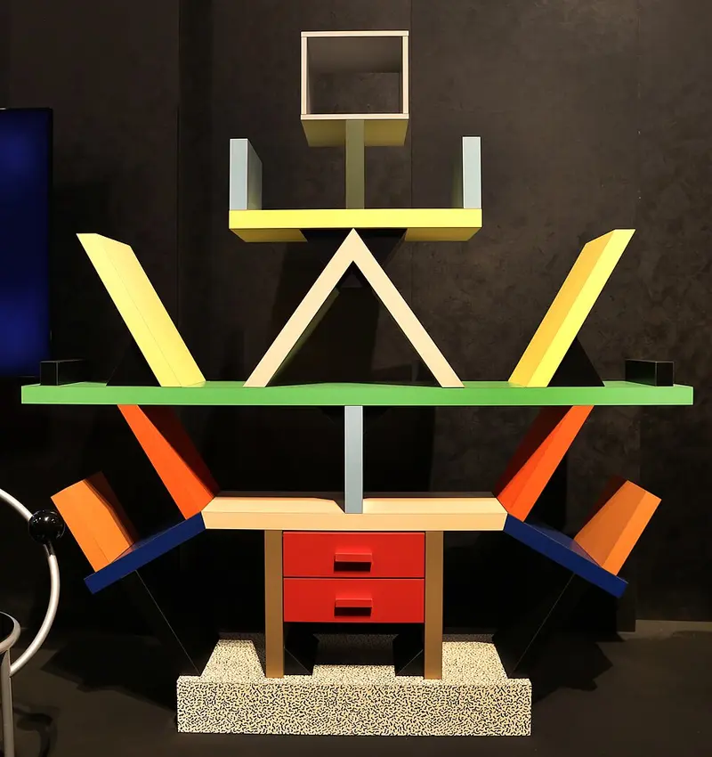 A colourful room divider like a totem, with shelves sticking out at odd angles in yellow, green, orange, red and blue, on a base covered in a black and white squiggle pattern.