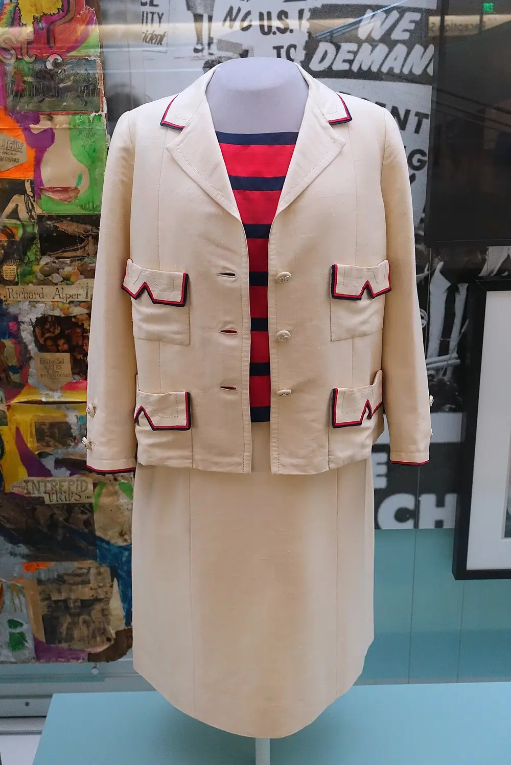 A cream three-piece suit by Chanel on a museum mannequin: a straight, collared jacket with four pockets trimmed in red and navy, a red and navy striped top, and a straight skirt.