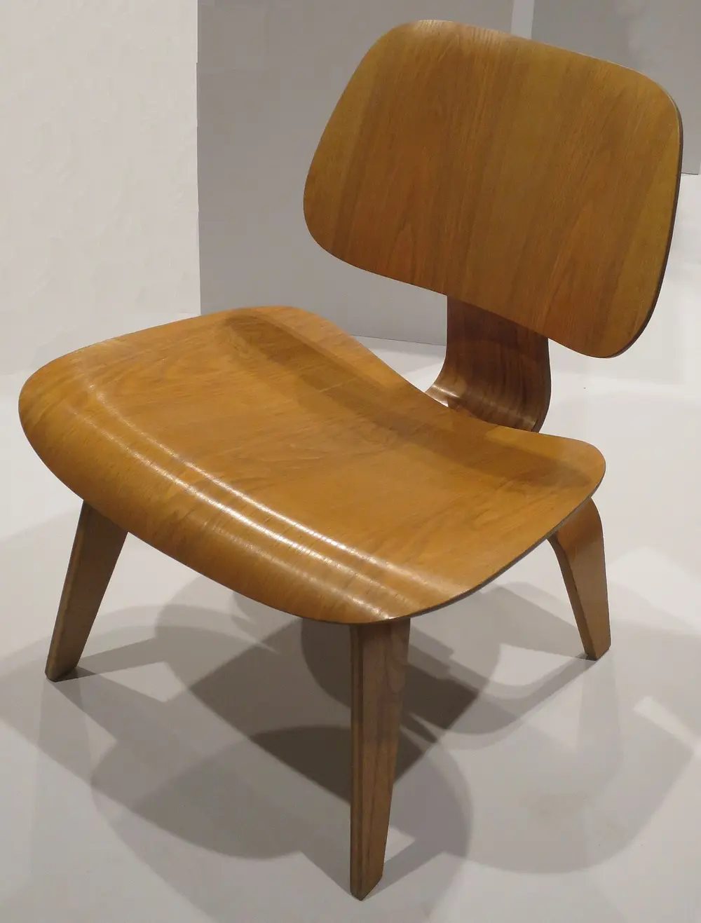 A low chair made of two curved panels of golden plywood, a seat and a back, joined by a bent plywood spine and standing on four plywood legs.