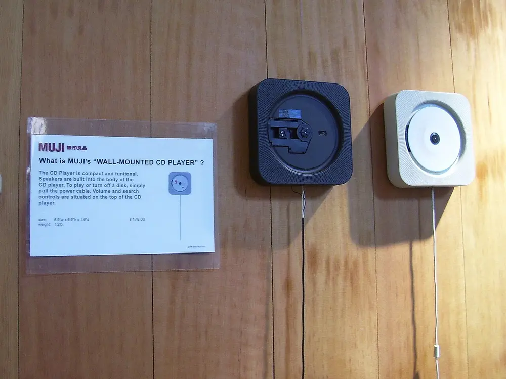 Two square CD players hanging on a wooden wall in a shop, one white and one black, each with a long cord hanging down, next to a Muji sign explaining the wall-mounted CD player.