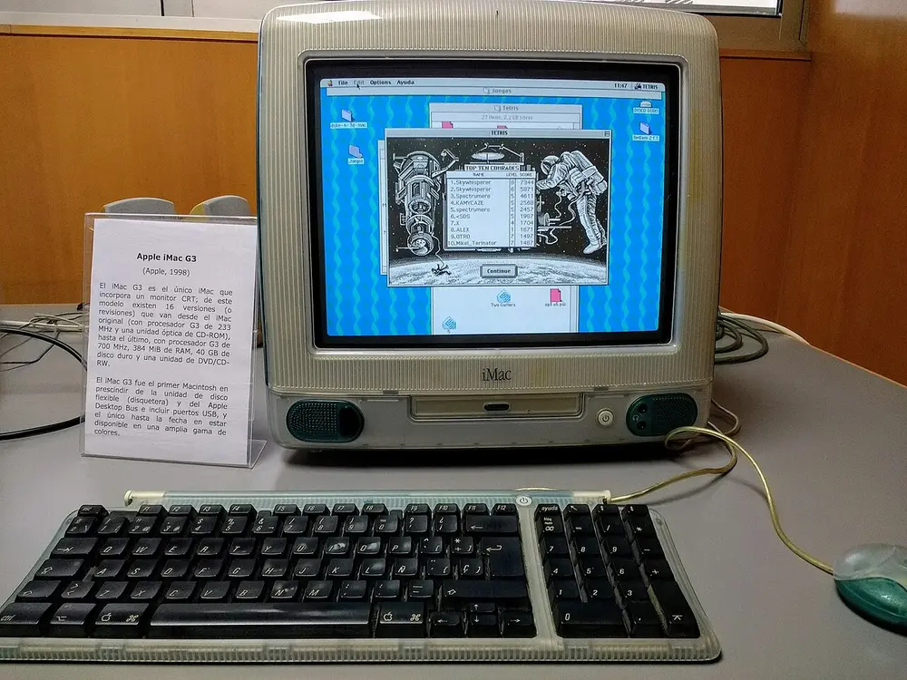 An egg-shaped all-in-one computer of see-through blue-green and white plastic with a curved screen showing a game, a matching see-through keyboard and a round mouse, on a museum desk beside a label.