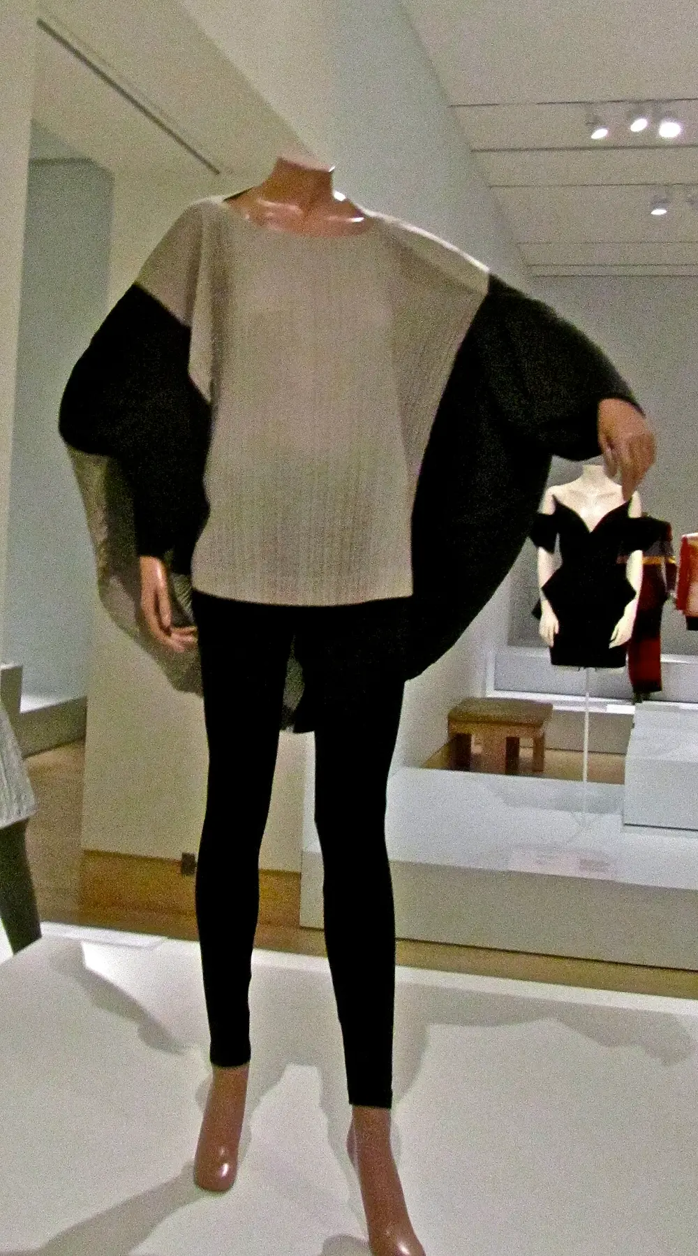 A loose, finely pleated top in grey with huge rounded black and silver sleeves that curve out like a circle, worn over black leggings on a mannequin in a museum.