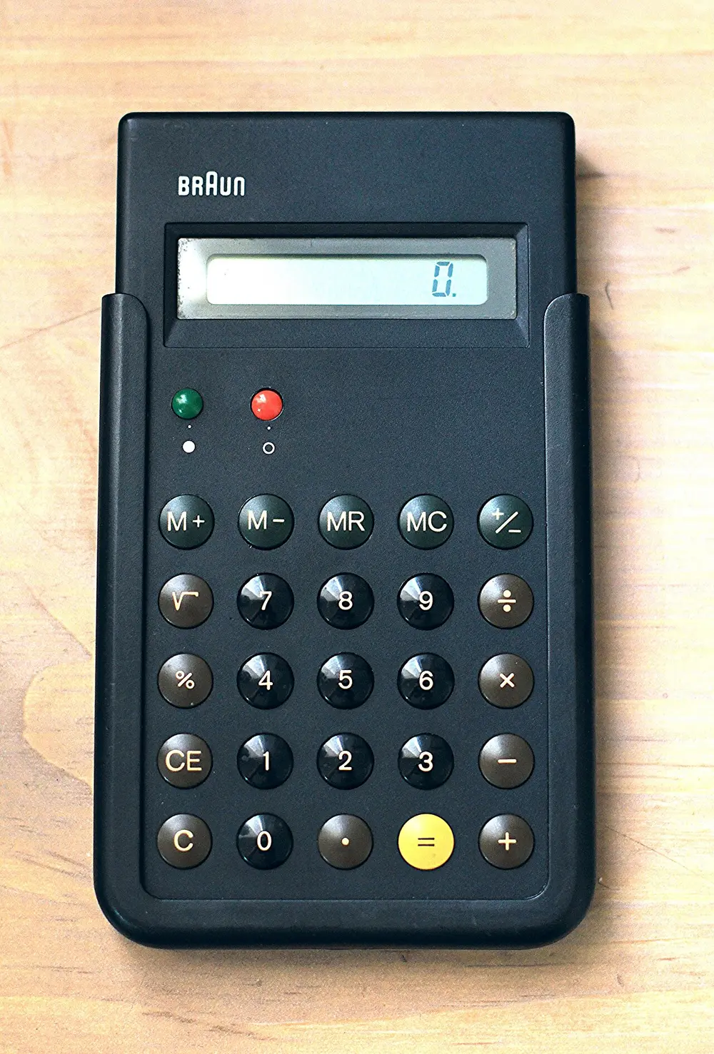 A slim black pocket calculator marked Braun, with a small grey display and rows of round, domed buttons, a yellow equals key, and red and green dots near the top.