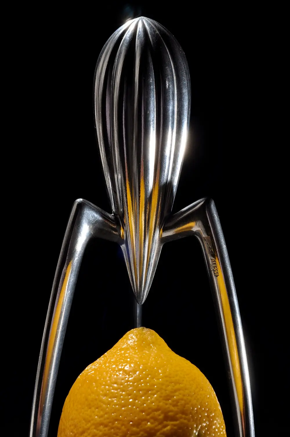 A tall, shiny aluminium lemon squeezer shaped like a ribbed teardrop on three long thin legs, with a lemon placed below it, against a black background.