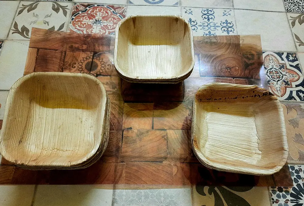 Three square brown plates pressed from dried palm leaf, with a visible natural grain, on a patterned tiled floor.