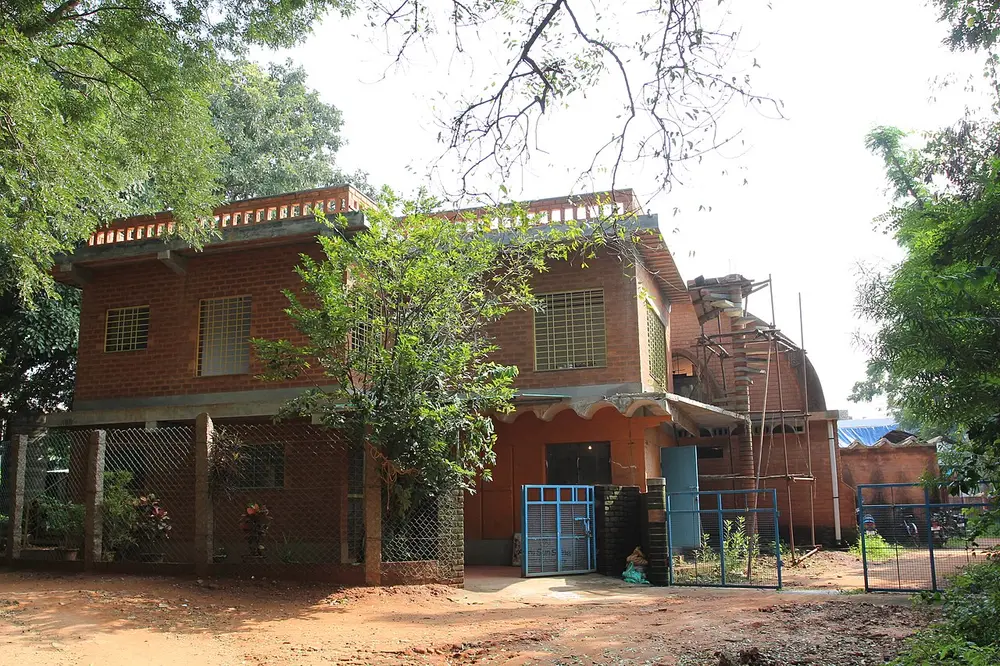 A two-storey building of neat orange-brown earth blocks behind trees, with a curved vault of the same blocks being built on one side under bamboo scaffolding.