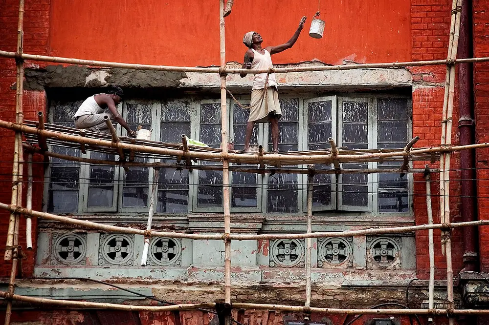 Two workers painting an old red building in Kolkata while standing on scaffolding made of bamboo poles tied together with rope.