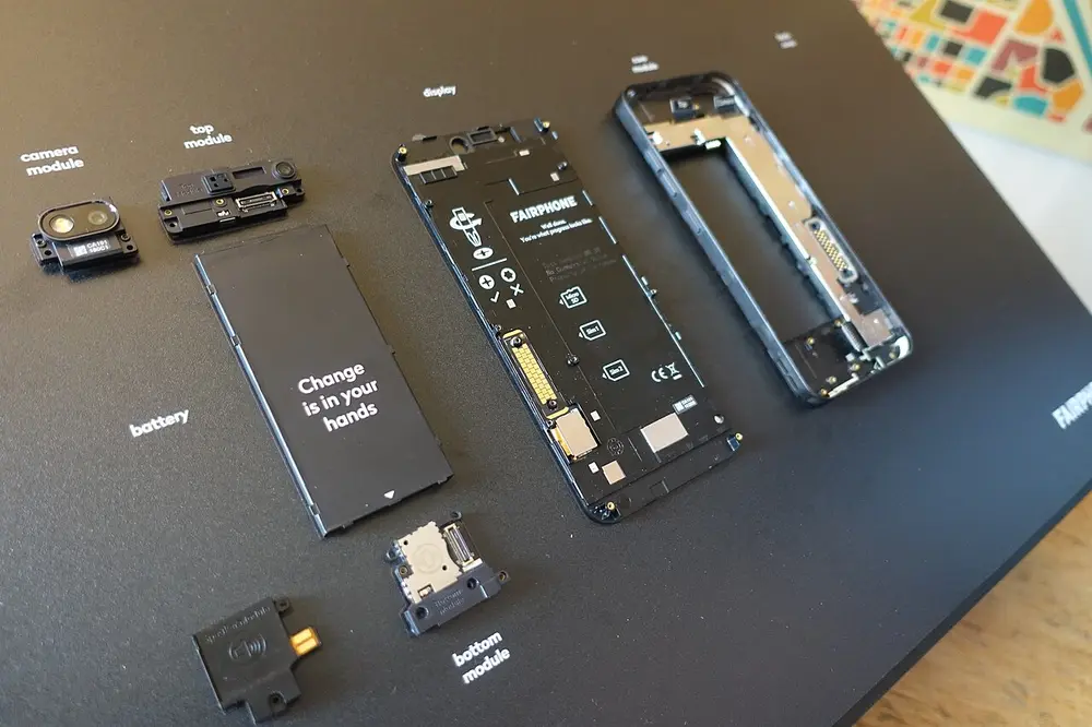 The parts of a smartphone laid out on a black board and labelled: the battery, the screen, the frame, the camera and other small modules.