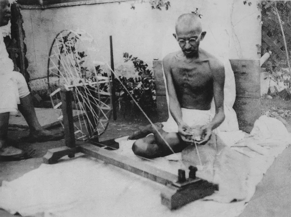 A black and white photo of Mahatma Gandhi sitting on the ground, spinning thread on a wooden spinning wheel.