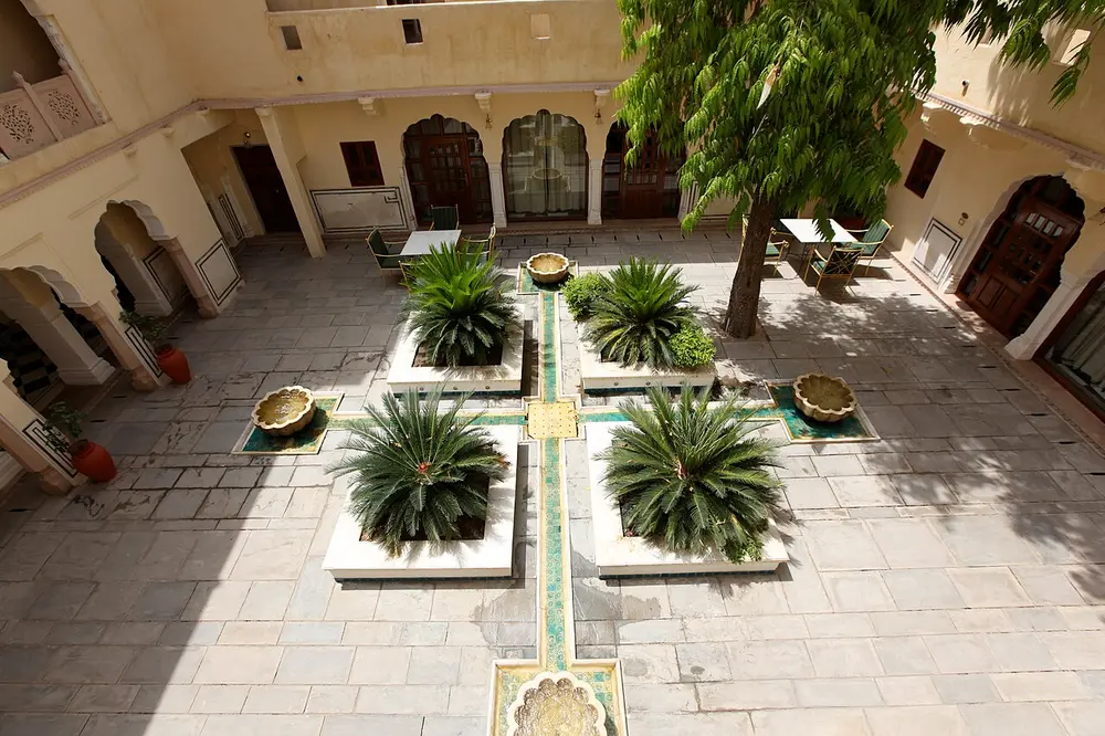 A paved courtyard seen from above, with four small gardens of palms and a cross of green tiles, surrounded by cream walls with arched doors and windows.