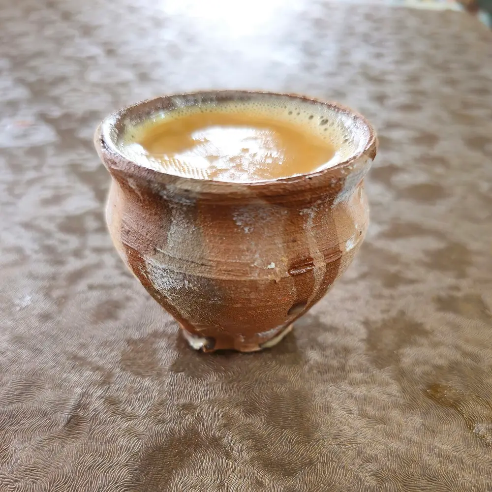 A small rough brown clay cup full of milky tea, on a table.