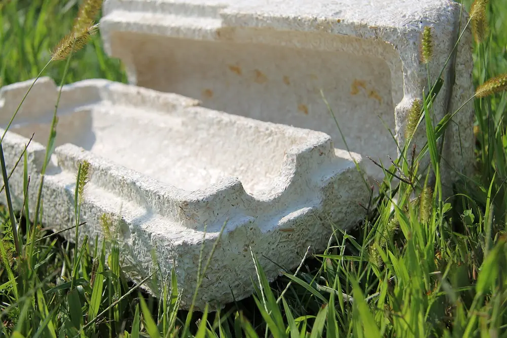 A white foam-like packaging box with moulded slots, lying in long grass, made from mushroom roots grown around farm waste.