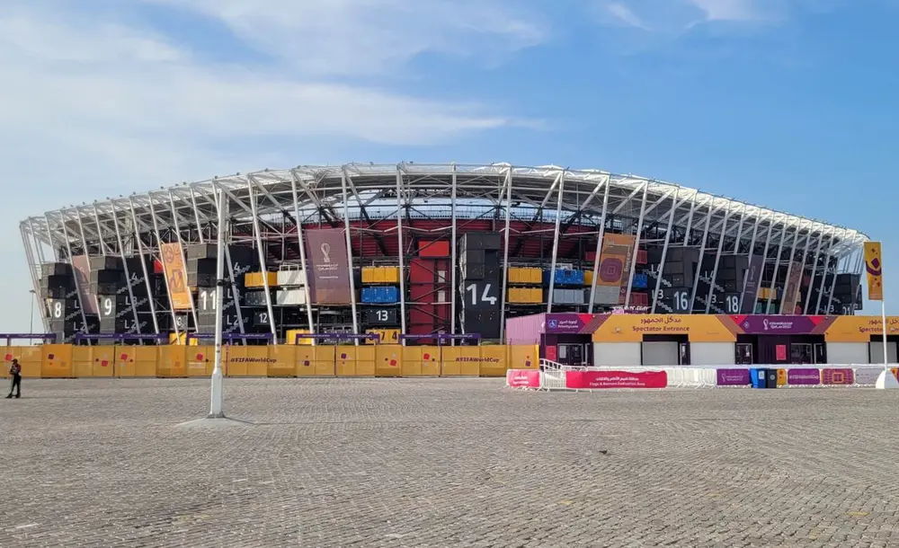 A football stadium whose walls are made of stacked shipping containers in red, blue, yellow and grey, held in a white steel frame, under a blue sky.