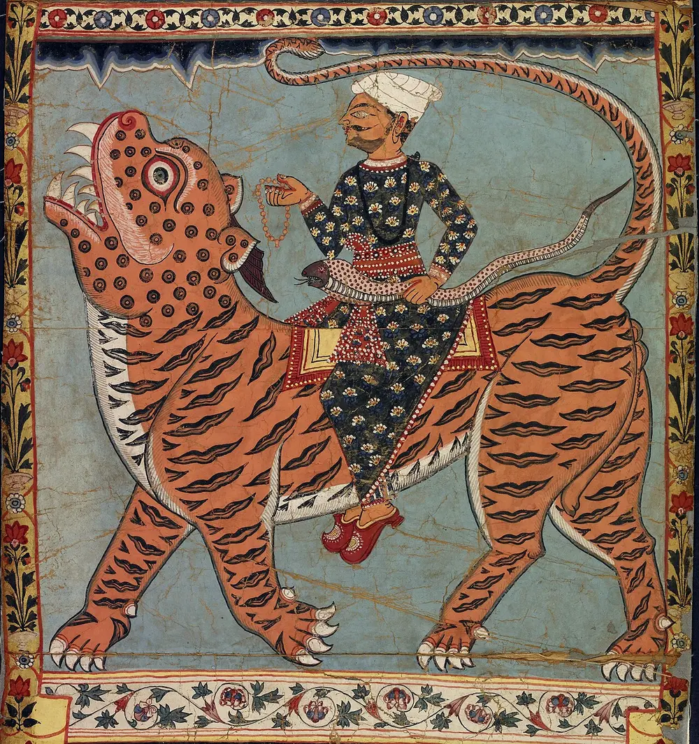 A man in a turban sits cross-legged on the back of a huge striped tiger with a spotted face, on a blue ground inside a flowered border.
