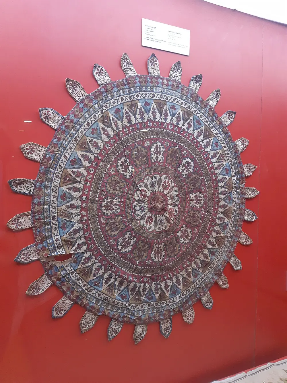 A round cloth printed with rings of fine flower patterns in red and blue, displayed on a red museum wall.
