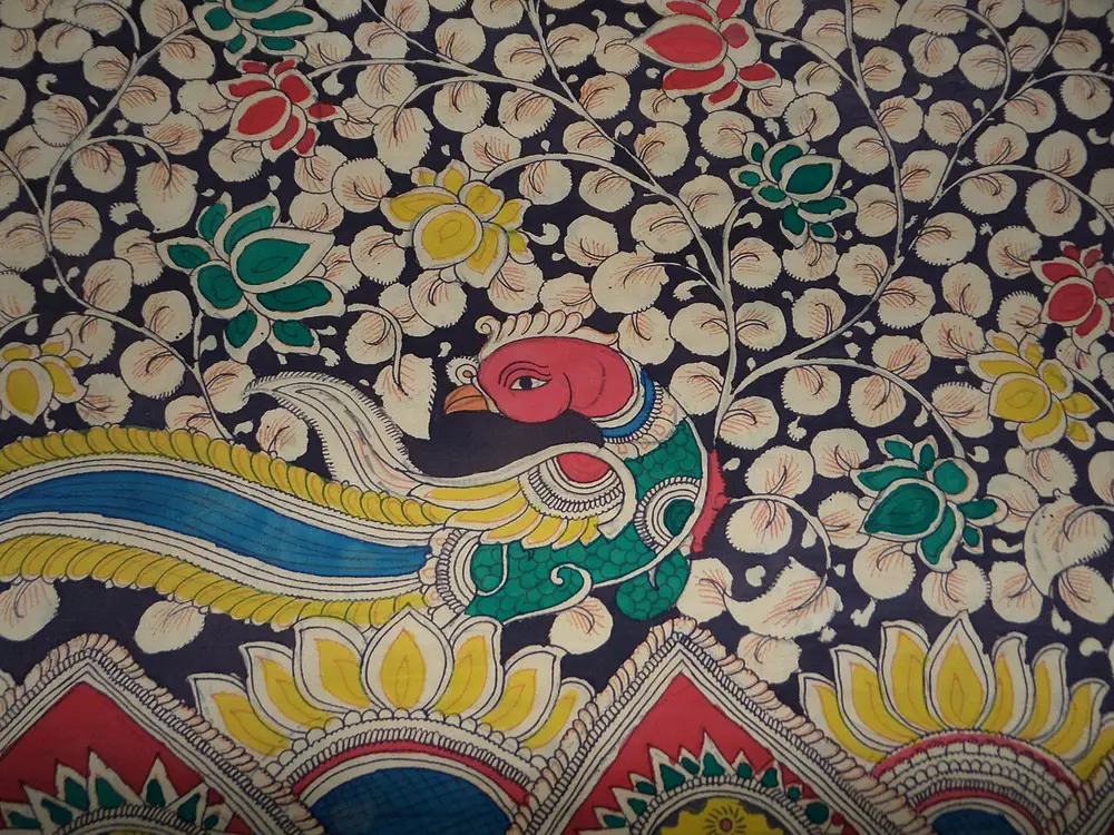 Part of a hand-painted cloth from Srikalahasti showing a peacock with a long blue and yellow tail among lotus flowers and pale leaves on a dark ground.