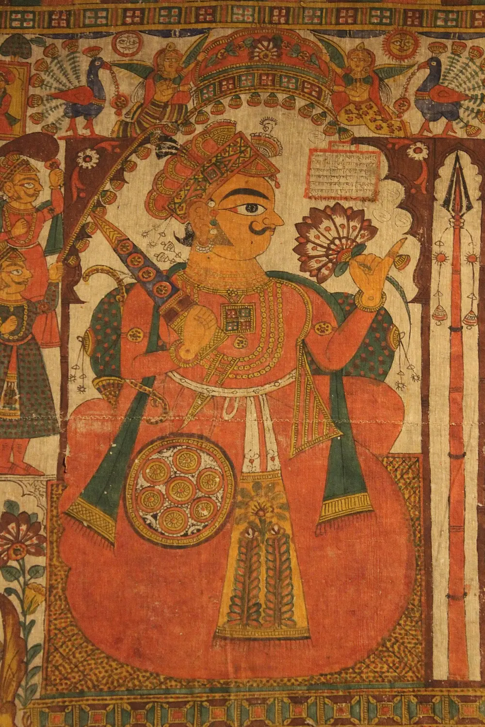 A detail of a painted cloth showing a large hero figure with a curled moustache, a shield and a spear, seated under an arch, painted mainly in orange-red and yellow.