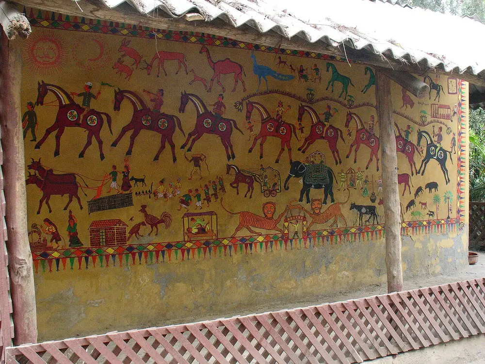 The outside wall of a hut painted with rows of colourful horses with riders, elephants, bullock carts and small figures on a pale yellow ground.