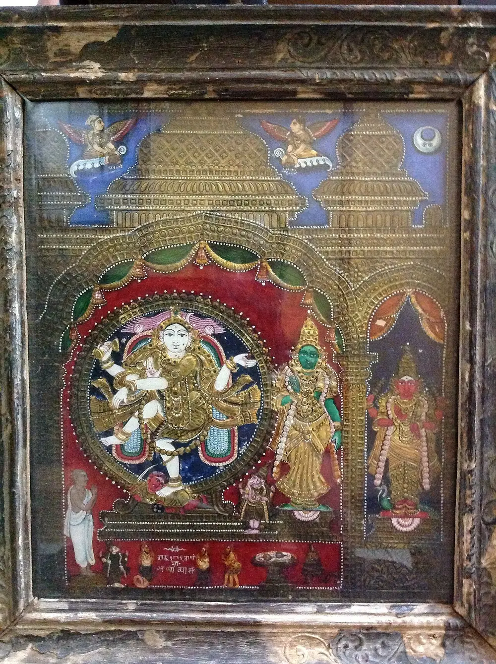 An old framed painting of the dancing Shiva, Nataraja, inside a ring, with his consort beside him, under raised golden arches studded with small coloured stones.