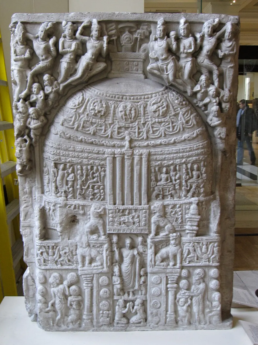 A pale limestone slab carved with a stupa covered in garlands and small scenes, with lions on pillars, the Buddha standing in the gateway and crowds of flying figures above.