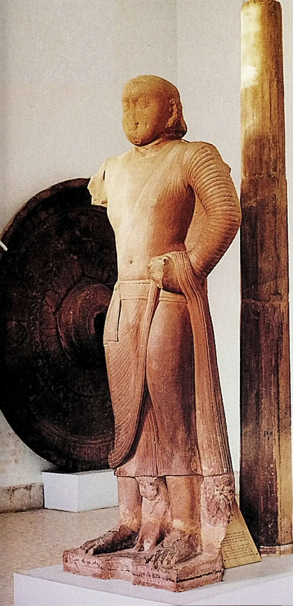 A tall standing red sandstone figure with a round face and a shaven head, his robe gathered at the waist, with a large carved stone umbrella and its tall shaft displayed beside him.