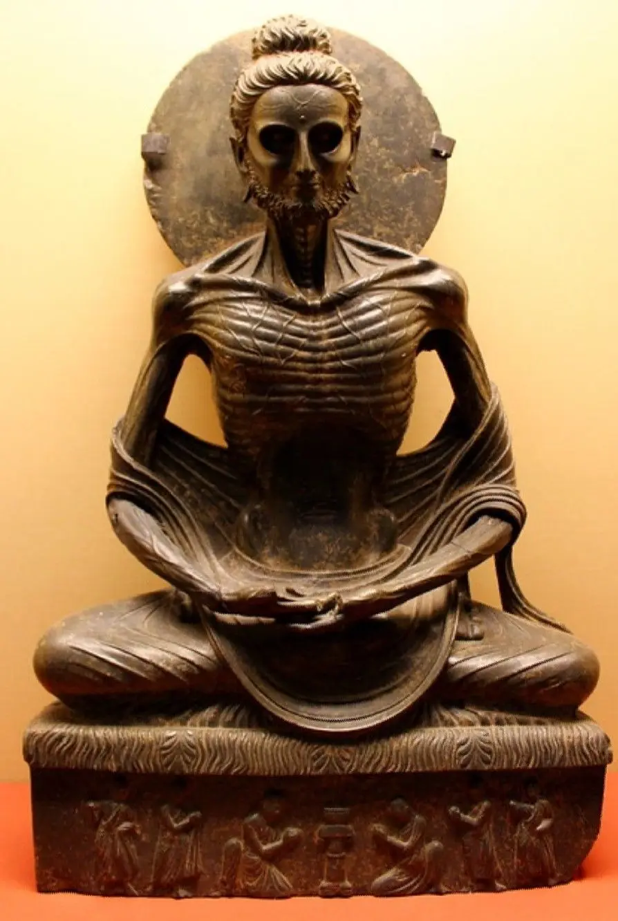 A dark grey stone figure of a seated man so thin that every rib and vein shows, with sunken eyes, a beard and a round halo behind his head.