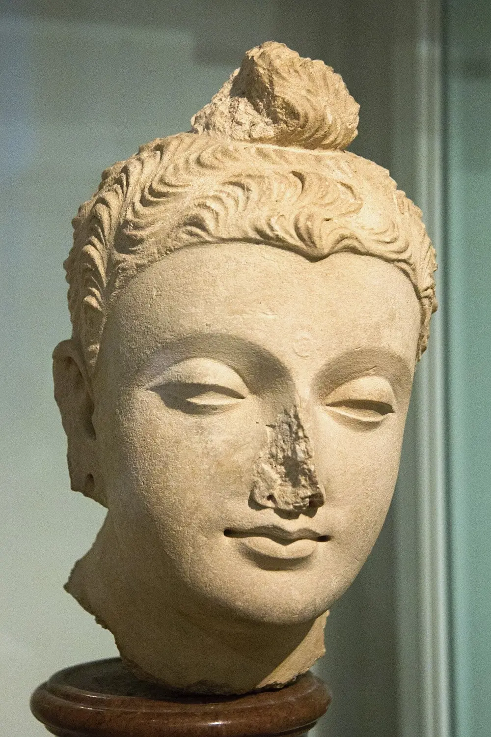 A pale cream plaster head of the Buddha with deep wavy hair, a high topknot, half-closed eyes and a broken nose.