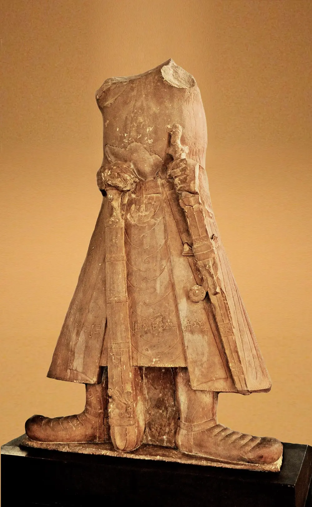 A headless red sandstone statue of a king in a long stiff coat and heavy padded boots, holding a sword at his side and a large mace.