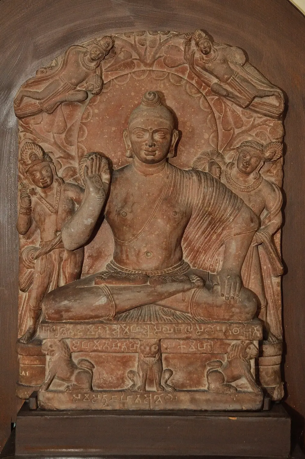 A red sandstone panel of the Buddha seated cross-legged on a lion throne, right hand raised, with a large halo, two standing attendants and two flying figures above.