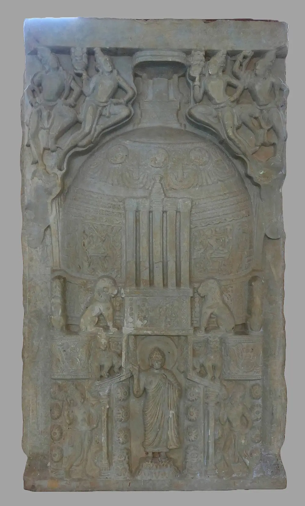A grey-white limestone slab carved with a large domed stupa, two lions at its base and a standing Buddha with a halo in the gateway, with flying figures at the top.