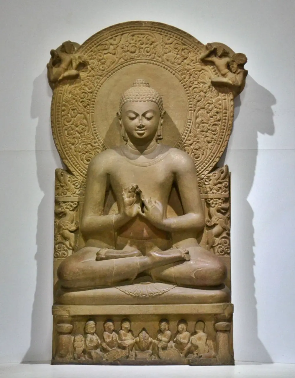 A buff sandstone Buddha seated cross-legged with his hands held at his chest, eyes lowered, in front of a huge round halo carved with flowers and creepers; small figures and a wheel are carved on the base.