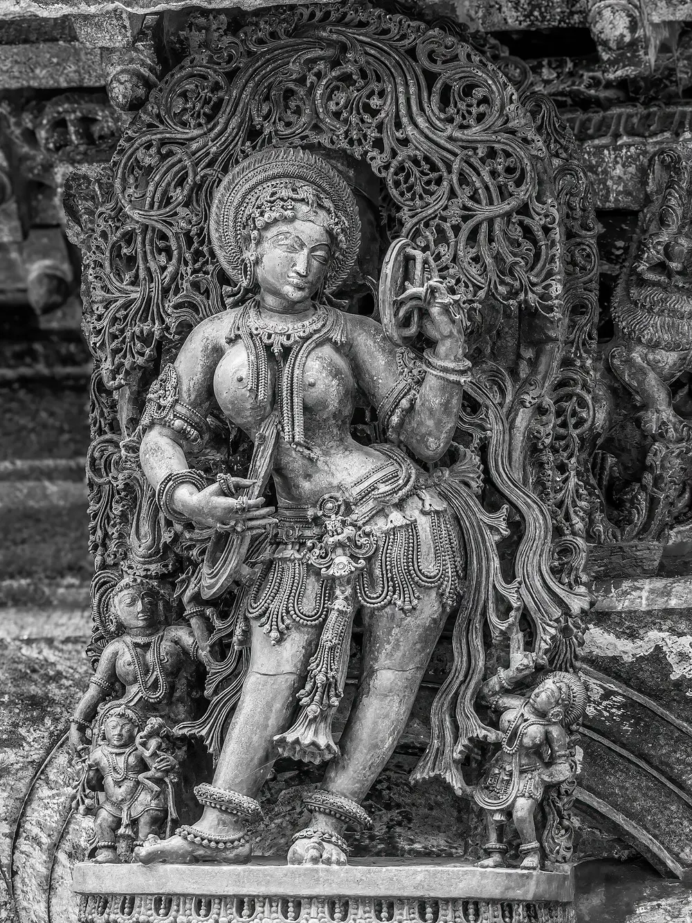 A black-and-white close view of a stone figure of a woman holding up a mirror and looking into it, covered in carved jewellery, under an arch of very fine lacy foliage; small figures stand at her feet.