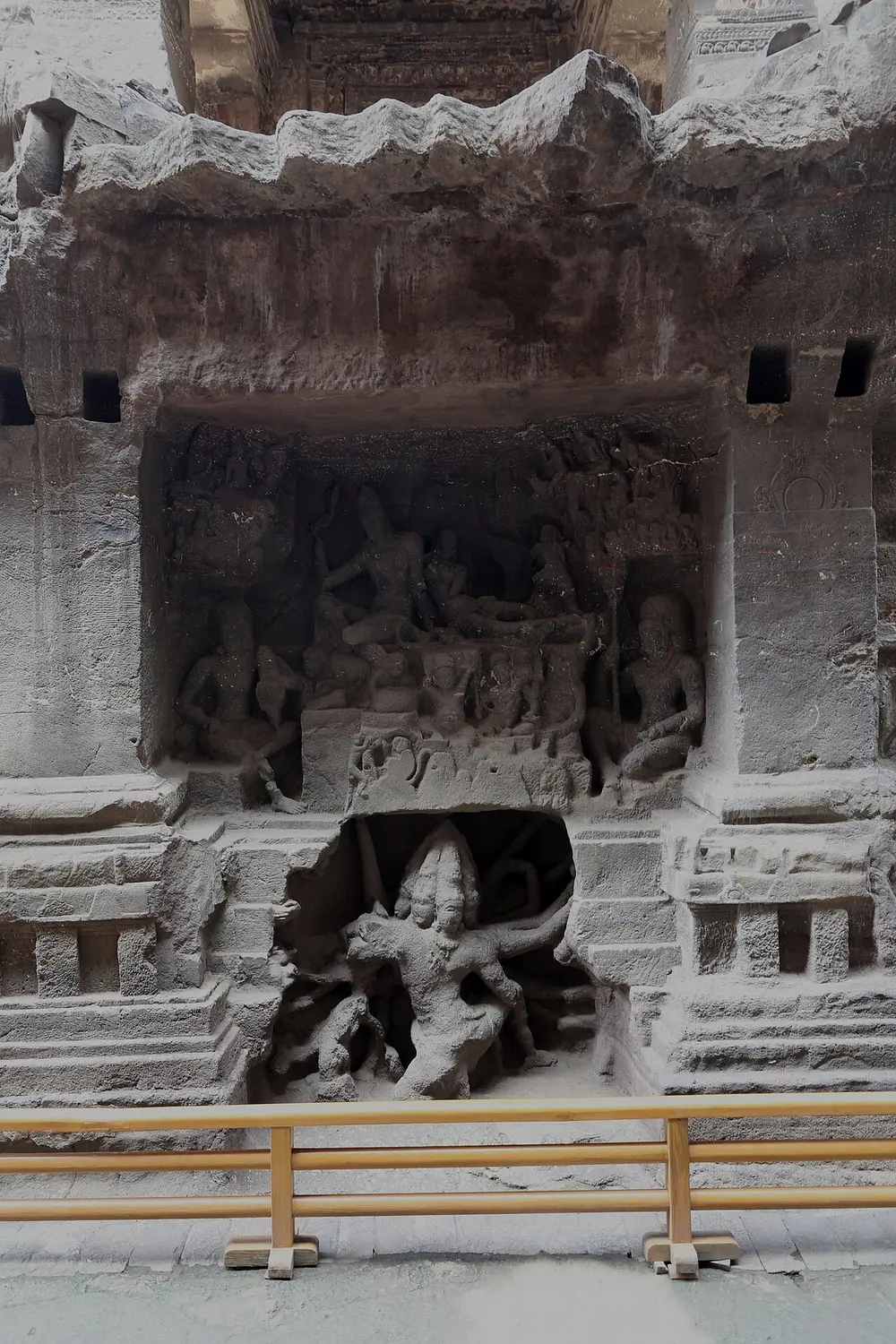 A deep relief cut into the side of a dark stone temple: above, Shiva and Parvati sit on a mountain with attendants; below, in a cave-like hollow, a many-headed, many-armed figure strains to lift the mountain.