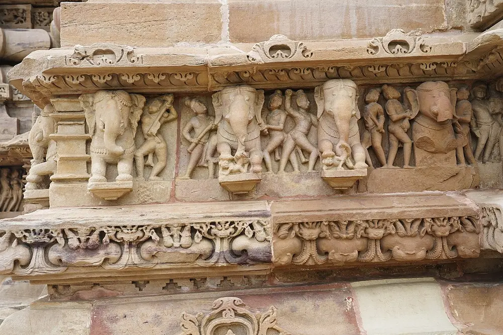 A long band of pale sandstone carving on a temple base: elephants face outwards between rows of soldiers, musicians and dancers, with bands of carved patterns above and below.