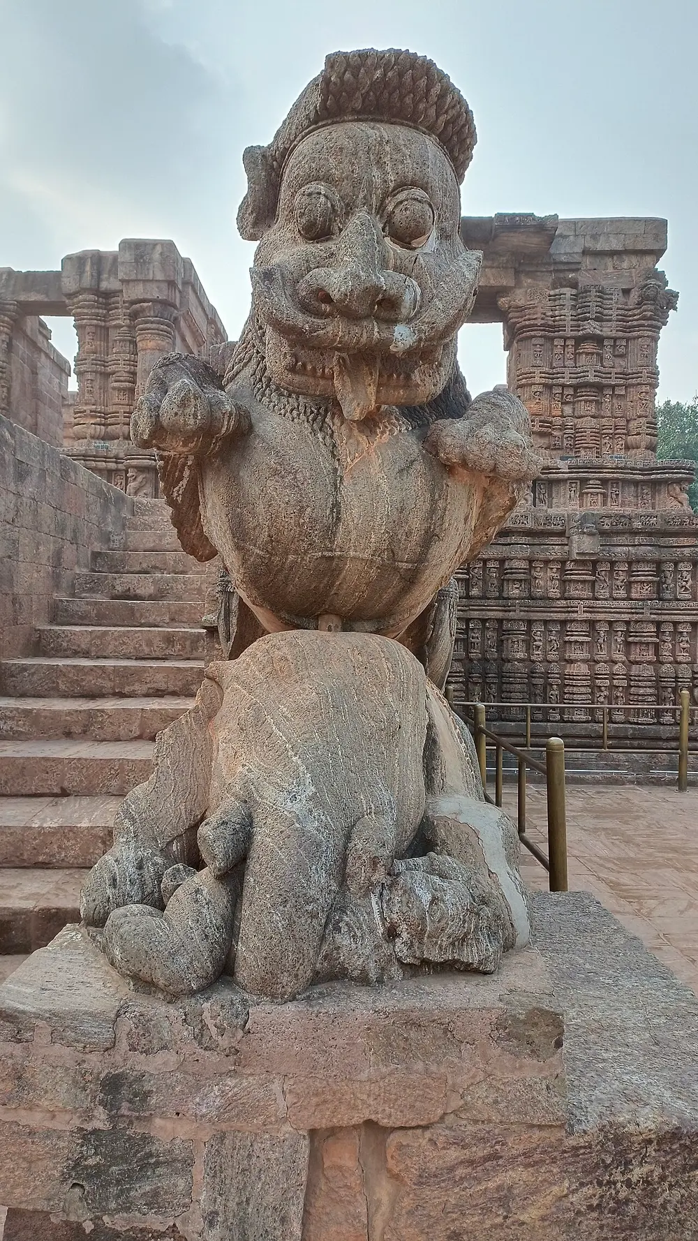 A large weathered stone lion with bulging eyes and a lolling tongue, rearing up on top of a stone elephant, at the foot of a flight of steps, with a carved temple hall behind.